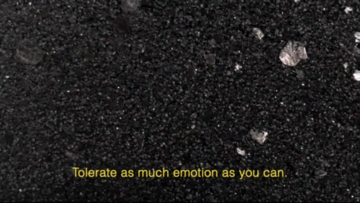 Description: [A flat screen monitor sits on the floor leaned against a gallery wall. The image on screen is of a rocky asphalt ground. Captions on the video image read: "Tolerate as much emotion as you can".]
Photo credit: Jonathan Dorado

Park McArthur and Constantina Zavitsanos, *Scores for Carolyn*, 2019
HD video, open captions, slowed sound. 11 min 28 sec  - © The artists and Galerie Max Mayer, Paris Internationale